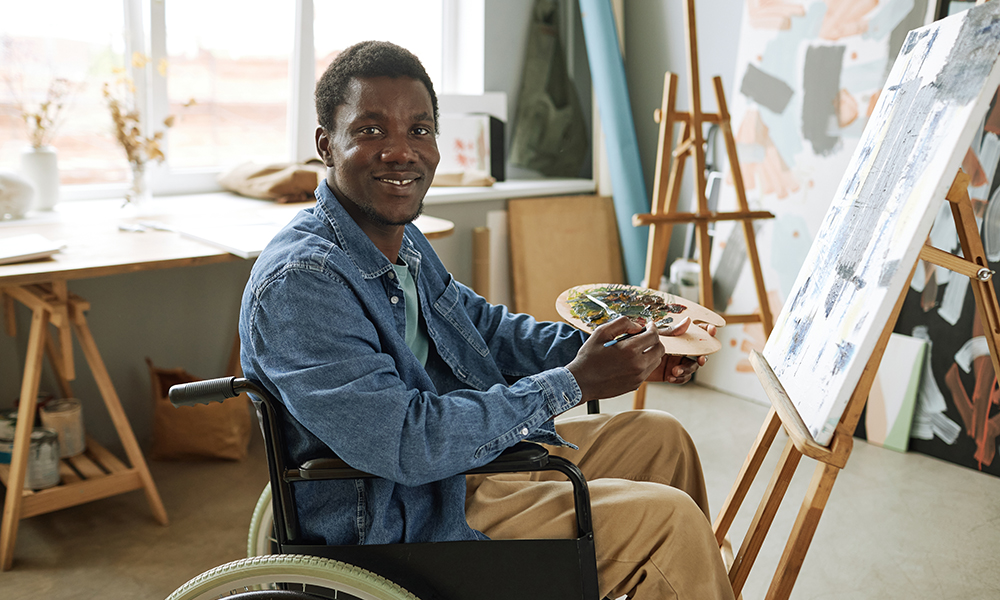 young man sits in front of a canvas with a palette and paintbrush in a wheelchair